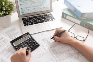 tributação NCM - homem utilizando uma calculadora enquanto preenche vários documentos expostos em cima de uma mesa com um notebook. Title content: Entenda como funciona a tributação NCM