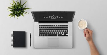 4 razões para automatizar a gestão fiscal e tributária do seu negócio - Mesa com notebook, caderno de anotações e um braço segurando uma xícara de café